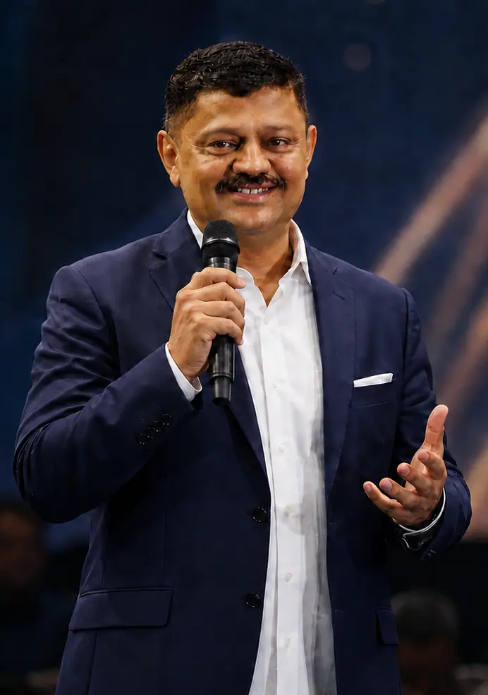 Satyanarayana Sheshagiri speaking on stage with a microphone, mid-gesture